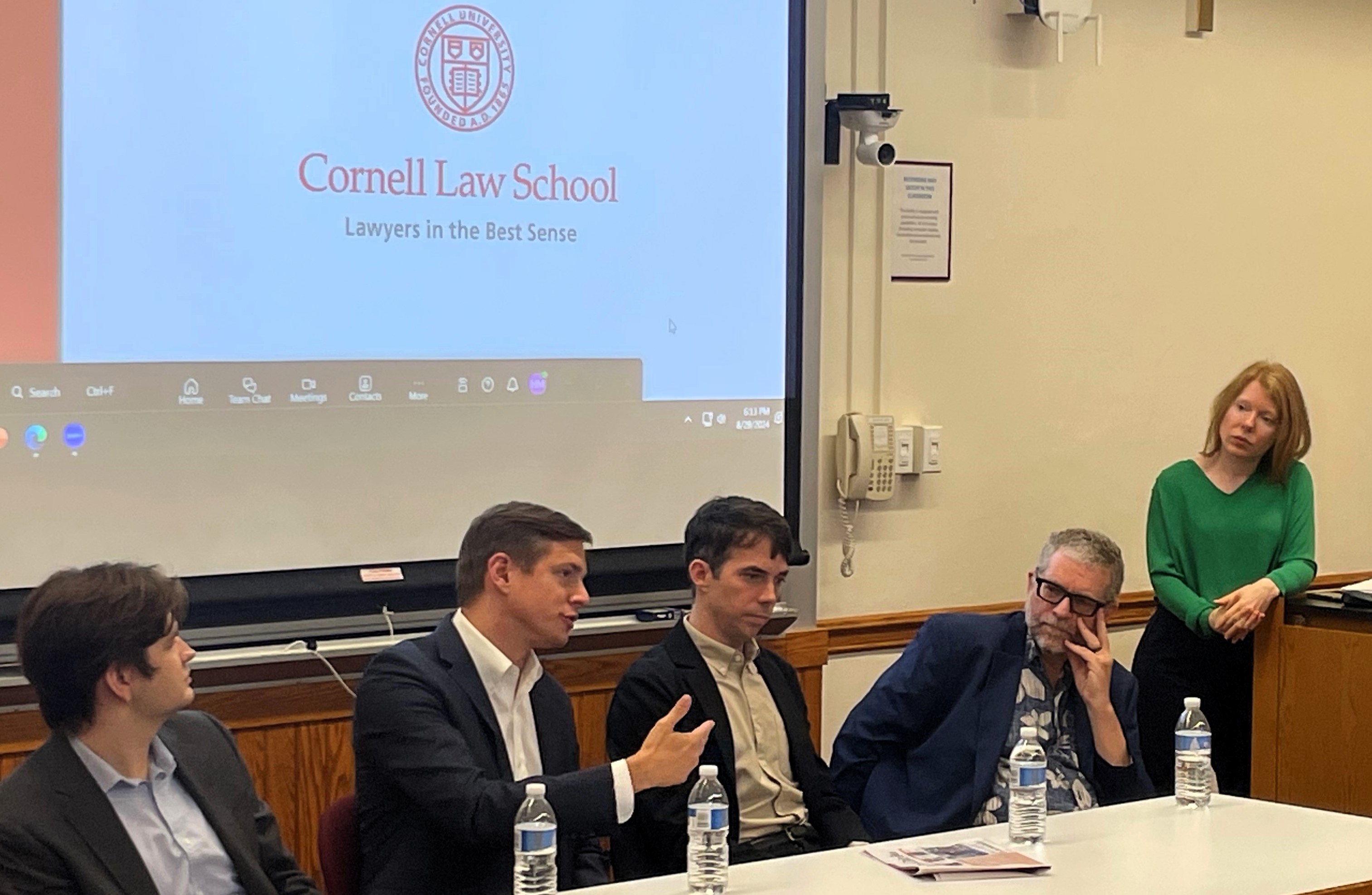 panel of four mean present in front of a screen with a Cornell Law School logo on it while a female moderator looks on