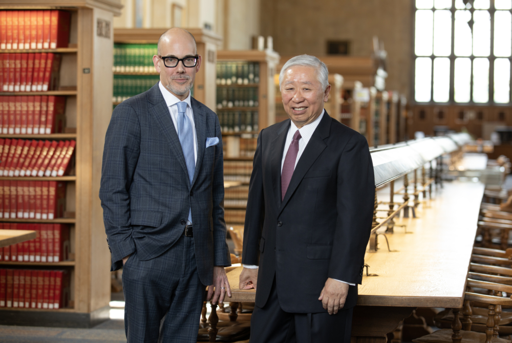 Jonathan Zhu, standing in front of a table in the law school library, wearing a black jacket and red tie and Dean Jens Ohlin next to him in a gray jacket and blue tie