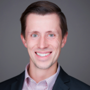 Austin Arnold smiling wearing a checkered shirt and suit jacket on a gray background.